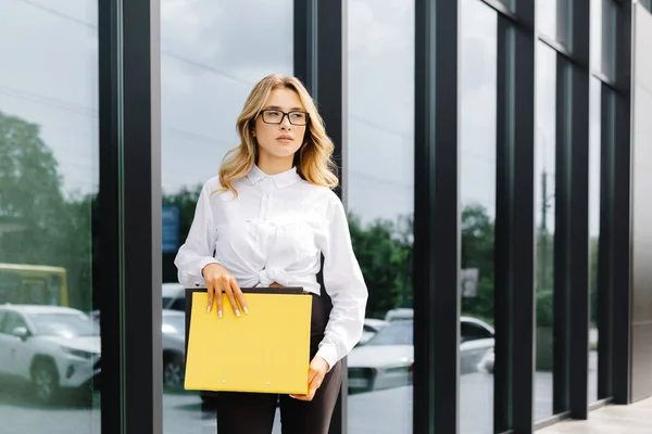 エレガントな自信を持った若い女性は 正式に服を着て ビジネスセンターの背景に対して屋外に立って 文書付きのフォルダを保持し カメラを見ます — ストック写真