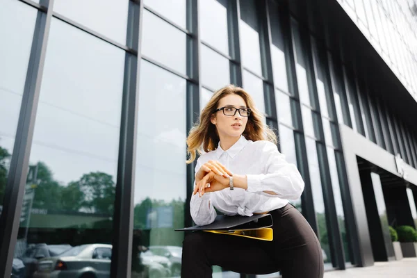 エレガントな自信を持った若い女性は 正式に服を着て ビジネスセンターの背景に対して屋外に立って 文書付きのフォルダを保持し 時計を見ます — ストック写真