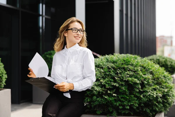 エレガントな自信を持った若い女性は 正式に服を着て ビジネスセンターの背景に屋外に座り 文書付きのフォルダを保持し 時計を見ます — ストック写真