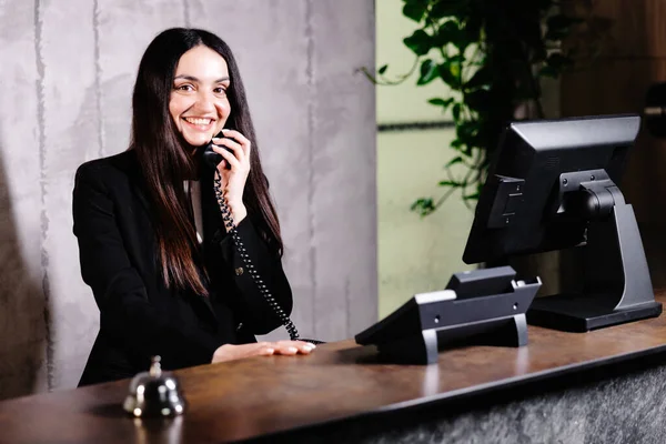 Hotel receptionist. Modern hotel reception desk with bell. Happy female receptionist worker standing at hotel counter and talking to phone