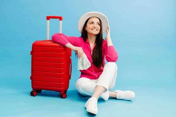 stock image Traveler tourist woman in casual clothes and white hat holding money and sitting at the red suitcase isolated on blue background. Female passenger traveling abroad to travel weekends getaway.