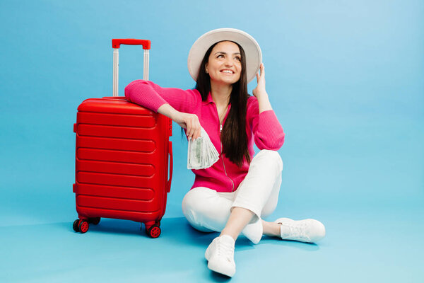 Traveler tourist woman in casual clothes and white hat holding money and sitting at the red suitcase isolated on blue background. Female passenger traveling abroad to travel weekends getaway.