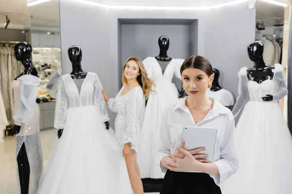Attractive bridal shop owner women use digital tablet help customer choosing wedding gown at the store.