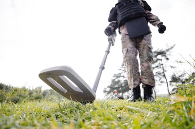 A man in a military uniform and bulletproof vest works in the forest with a metal detector. A minesweeper performs work on demining the territory. clipart