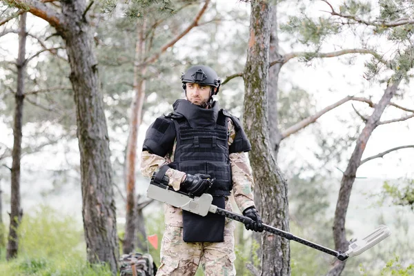 stock image A man in a military uniform and bulletproof vest works in the forest with a metal detector. A minesweeper performs work on demining the territory.