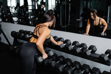 Spor giyim bölümündeki güzel kadın aynaya bakarken askıya yaslanıyor ve spor salonunda ağır halterleri kaldırıyor..