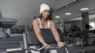 Genç bir kadın halterli ve kavisli bir halterle kolları için egzersiz yapıyor, bankta oturuyor. Spor salonundaki kadın beyaz takım elbise giyiyor ve spor yapıyor.