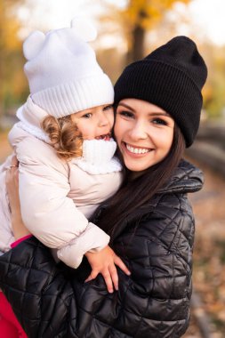 Sarı saçlı anne ve küçük kız, bir sonbahar parkında sarı ve turuncu yapraklı arka planda oynuyorlar. Aile parkta yürüyor.