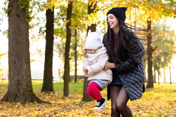 Mother and young little girl with blonde hair are playing in an autumn park on a yellow and orange leaf background. Family is walking in park