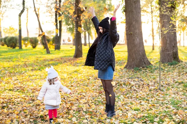 Mother and young little girl with blonde hair are playing in an autumn park on a yellow and orange leaf background. Family is walking in park