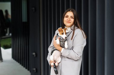 Sıradan bir kadın, en sevdiği tüylü, eğitimli, saf Jack Russell Terrier köpeğine sarılıyor sonbahar günü açık havada, duvarın yanında köpekler için takım elbise giyiyor. Sadık en iyi arkadaşlar