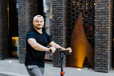 Young handsome man in white baseball cap riding on scooter to work along the street in the city. Active lifestyle, backpacker traveler. Ecology and clean ecological mode of transport