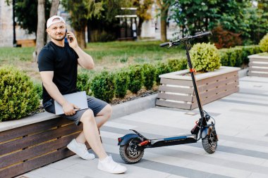 Elinde dizüstü bilgisayar olan yakışıklı serbest çalışan ve konuşan biri sokakta elektrikli scooter 'ın yanında oturuyor.