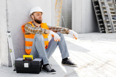 Tamirci, miğfer ve yelek giyen ustabaşı iş yerinde alet çantası ve cetvelin yanındaki binada dinlenmek için oturuyor..