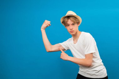 Pazılarıma bak. Sıradan beyaz tişörtlü, parlak mavi arka planda izole edilmiş, kol kaslarını gösteren komik gülümseyen genç bir çocuk resmi. Çocukluk, fitness ve spor konsepti