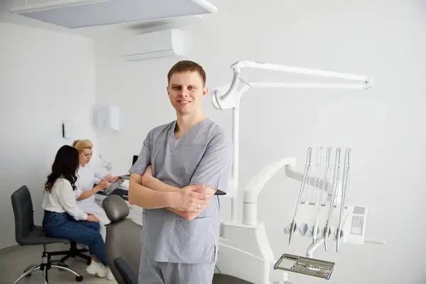A young male dentist stands in the middle of the office, and in the background a colleague is advising patients. Preparation for admission of patients