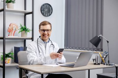 Male doctor medical worker in modern clinic wearing eyeglasses and white coat uniform using cell mobile smartphone apps, sitting at laptop computer. Medicine technologies health care concept