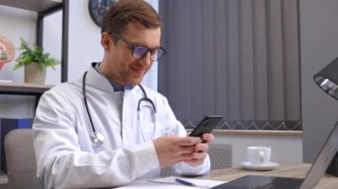 Modern klinikte çalışan, gözlüklü ve beyaz önlüklü bir erkek doktor cep telefonu uygulamaları kullanıyor, dizüstü bilgisayarın başında oturuyor. Tıp teknolojileri sağlık hizmetleri kavramı