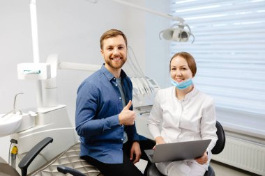 A handsome young man is talking to a female doctor at a dental appointment in a bright, beautiful office. The dentist explains to the patient and shows everything on the laptop.