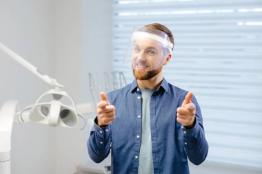 Portrait of handsome male dentist wearing in big protection medical shield on face preparing for dental surgery. Man smiling and looking at camera