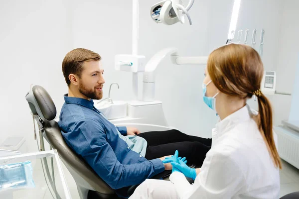 stock image Blonde female dentist in dental office talking with male patient and preparing for treatment. Handsome bearded man in dentist chair looking at his doctor with smile.