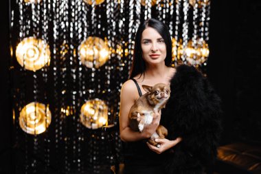 Young beautiful smiling brunette female in trendy evening clothes holding her litlle dog in her hands. Carefree woman posing near shiny tinsel wall in studio with dog. Fashionable model with bright makeup looks at camera.