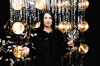 Happy woman with brunette long hair in trendy evening fur coat holding glass and confetti on shine wall in studio.