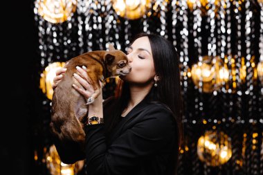 Young beautiful smiling brunette female in black jacket holding her litlle dog in her hands. Carefree woman posing near shiny tinsel wall in studio with dog