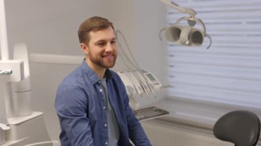 Genç sakallı adam dişlek gülüşünü işaret ediyor, ofisteki stomatoloji sandalyesinin yanında duruyor ve kameraya bakıyor.