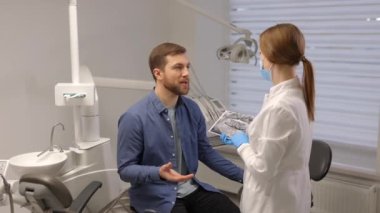 Young attractive man visiting dentist, sitting in dental chair at modern light clinic. Young woman dentist holding x ray image