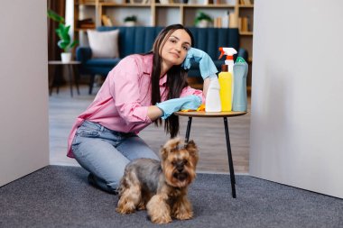 Evdeki temizlik malzemelerinin yanındaki masaya yaslanan genç bir kadın ve küçük köpeği onun yanında duruyor..