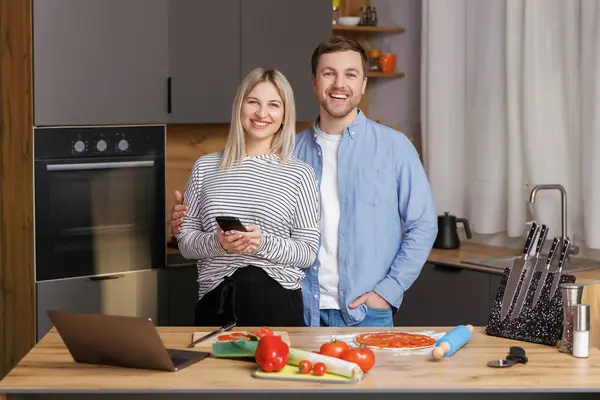Smiling loving couple cooking salad together while standing on a kitchen at home and using mobile phone. Cute couple man and woman taking photo of food and taking pictures together while cooking