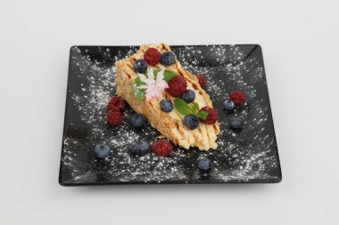 İştah açıcı bir Napolyon pastası. Böğürtlenlerle süslenmiş ve beyaz arka planda siyah bir tabakta nane şekeri..