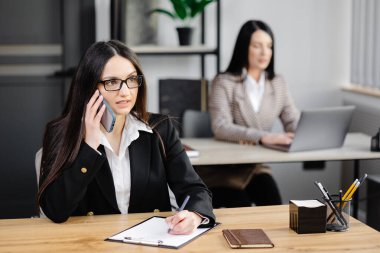 Çekici, ceketli genç bir iş kadını bir masada otururken cep telefonuyla konuşuyor ve ofisteki iş arkadaşının yanında not defteri kullanıyor..