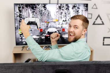 Genç adam evde vakit geçiriyor, kanepede oturuyor ve konsolda video oyunları oynuyor. Oyun oynamak ve kazanmak için joystick kullanan başarılı bir erkek..