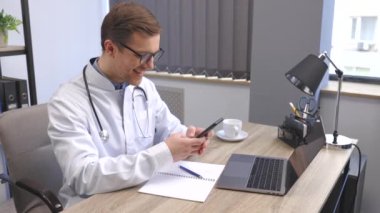 Male doctor medical worker in modern clinic wearing eyeglasses and white coat uniform using cell mobile smartphone apps, sitting at laptop computer. Medicine technologies health care concept.
