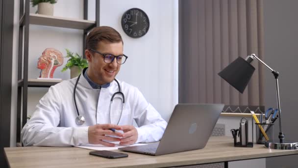 男医生在笔记本电脑屏幕上与在线病人交谈的肖像 坐在诊所的写字台上 为家庭健康治疗提供在线咨询 远程医疗预约 — 图库视频影像