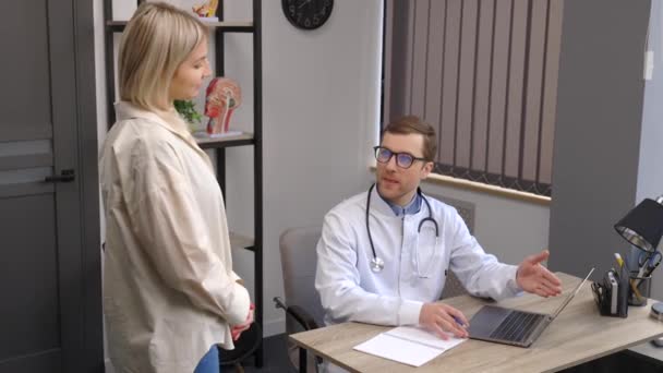 一位年轻貌美的医生正在他的办公室里进行会诊 医生正在笔记本电脑上做笔记 病人站在桌子旁边笑着 — 图库视频影像
