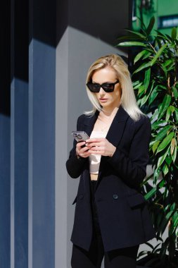 Siyah takım elbiseli genç iş kadınları arka plandaki gri duvarda izole edilmiş akıllı telefon kullanıyorlar. Güzel yönetici cep telefonu aygıtı ve mesaj tutuyor.