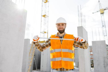 Tamirci, koruyucu miğfer ve yelek giyen ustabaşı inşaat alanında duruyor ve cetveli elinde tutuyor..