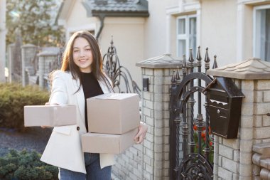 Kız evin yanında duruyor, elinde kutular ve kameranın önünde bir kutu tutuyor.