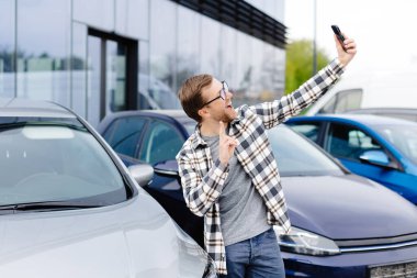 Mutlu adam yeni araba aldıktan sonra akıllı telefondan selfie çekiyor. Telefondaki video bağlantısıyla satın alınan arabayı gösteren adam..