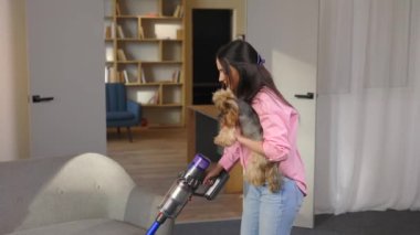 Elinde elektrikli süpürge ve köpeği olan bir kadın oturma odasını süpürüyor.