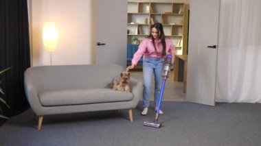 Sevimli genç bir kadın kablosuz elektrik süpürgesiyle evi süpürüyor ve küçük sevimli köpeğini eliyle okşuyor.