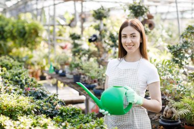 Genç bir kadın büyük bir seranın ortasında elinde bir su bidonuyla kameraya gülümsüyor.