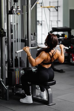 Siyah takım elbiseli kaslı atletik bayan vücut geliştirici spor salonunda spor simülatörüne çekildi. Sırt kasları çok gergin.