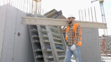 İnşaat sırasında inşaat alanında kereste taşıyan kasklı ve yelekli bir adam.