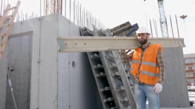 İnşaat sırasında inşaat alanında kereste taşıyan kasklı ve yelekli bir adam.