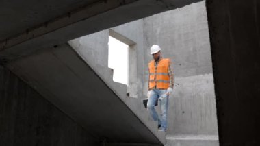 Tamirci, koruyucu miğfer ve yelek giyen ustabaşı inşaat alanında duruyor ve elinde cetvel ve alet çantası tutuyor.