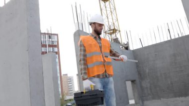 Tamirci, koruyucu miğfer ve yelek giyen ustabaşı inşaat alanında duruyor ve elinde cetvel ve alet çantası tutuyor.